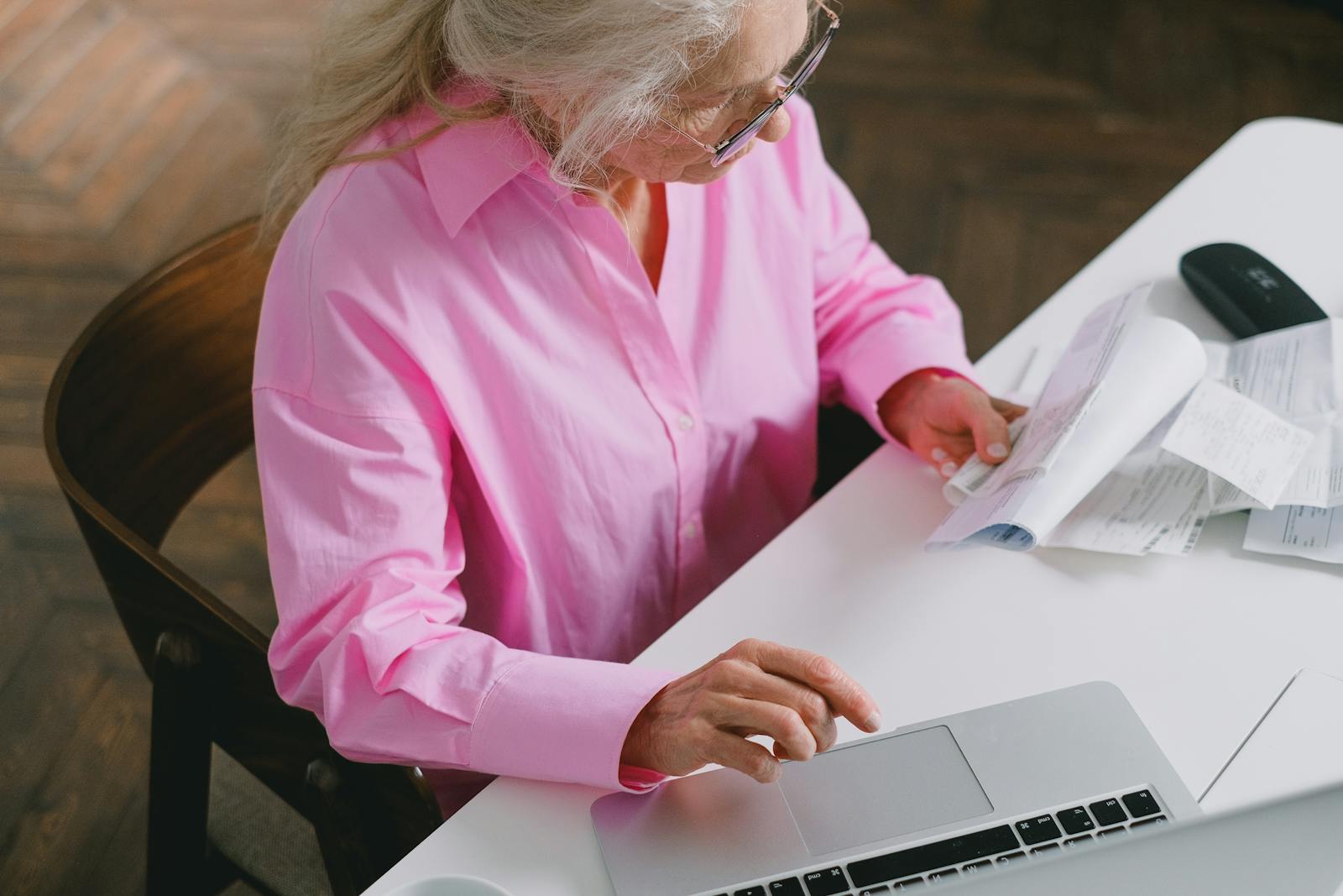 High-Angle Shot of an Elderly Woman in Pink Long Sleeves Looking at Documents while Using a Laptop